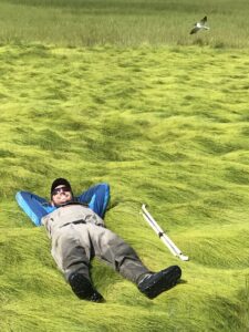 A person wearing sunglasses, a hat, and waterproof overalls smiling, laying down next to some white plastic poles in a field of marsh grass