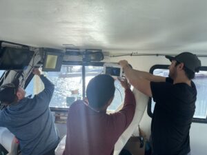 Three people inside the cabin of a boat connecting a small screen to the wall