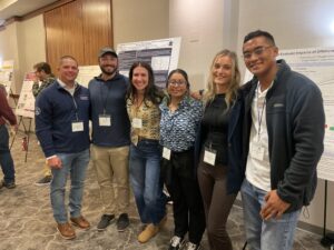 Six people wearing name tags posing for a picture with research posters and another person laughing in the background.